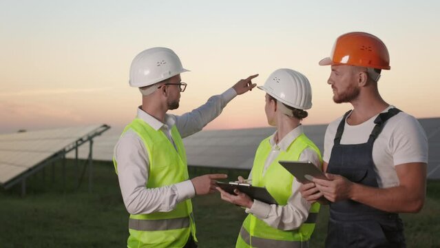 Male and female engineers with clipboard in hands talking with competent technician that using digital tablet. Outdoors meeting of coworkers among farm with solar cells.