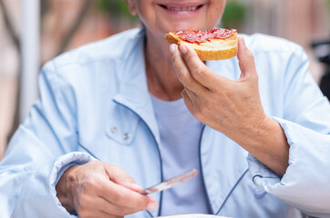 Mature Caucasian woman sitting at an outdoor table eating strawberry jam on toast. Healthy and...