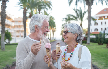 A nice elderly caucasian couple laughs while having fun eating the ice cream cone in the park. Joyful elderly lifestyle concept. Two white-haired seniors enjoying a walk