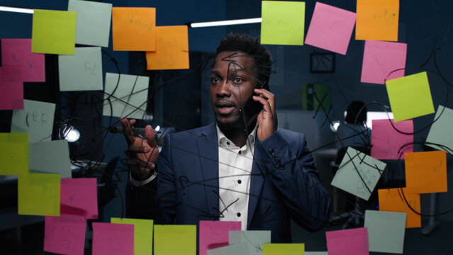 African-American businessman talk on cellphone looking at glassboard with notes in dark office