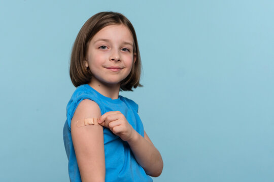 Child Vaccinated. Cute Little Girl pointing finger at adhesive plaster bandage on her arm after Being Injected Covid-19 Vaccine, light blue studio background. Children immunization. Health pass