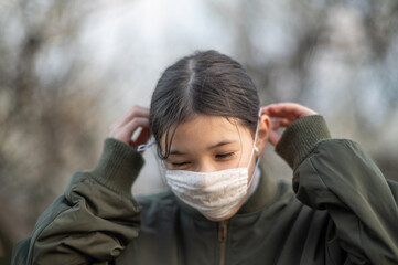 Young girl /child of mixed Asian -European ethnicity putting on a face mask to avoid virus / allergy. Close up outdoor shot during Covid-19.