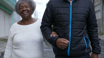 An aged son walking with elderly mother in 80s outside in urban street going for a walk