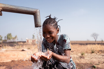 Happy little African girl crouching in front of a waterhole in an arid environment somewhere in the...