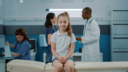 Obraz premium Portrait of little girl on bed waiting to begin checkup exam in medical cabinet with doctor. Small kid with illness looking at camera and getting ready for checkup visit appointment in office.
