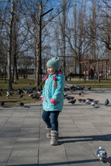 Fototapeta premium Preschool-aged girl in a blue jacket twirls around herself in front of a flock of city doves. Portrait of a five-year-old child with long pigtails holding her hands in her pockets. Sunny spring day.