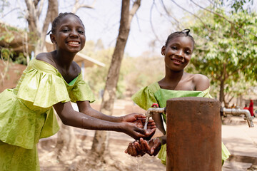 Two charming African girls in beautiful traditional dresses refresh themselves with water from a...