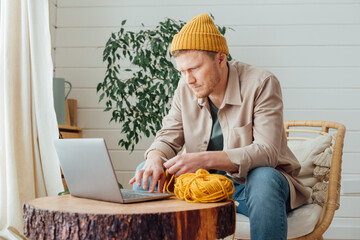 Young serious man looking at laptop. Man learning new hobby, knitting on needles. Knitting project in progress.