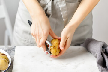 Obraz na płótnie Canvas Woman in apron peeling potato on marble board. White background. Hands peeling potato with kitchen peeler