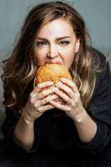 Hungry woman eats a hamburger. Beautiful blonde in gray pajamas. Junk food and unhealthy. Gray background. Vertical. Close-up.