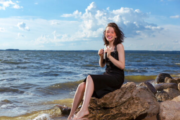 Positive Young Happy Smiling Caucasian Brunette Girl Posing in Black Dress On Stone At Sea During Sunny Day Outdoors.