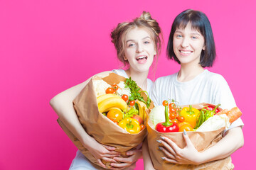 Couple of Two Positive Laughing Caucasian Girls Posing With Eco Paper Bags Filled With Grocery And Vegetables Over Pink Background.