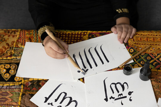 Close Up Muslim Girl Hands Writing Arabic Text With Bamboo Pens And Black Ink On Paper, Arabic Letters Mean The Name Of Muslim God 