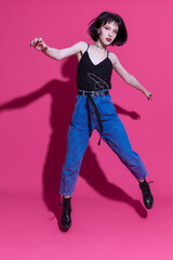 Fototapeta na wymiar One Happy Winsome Dancing Brunette Girl in Street Clothing Posing Blue Jeans and Bunch of Jewelry in Dance Pose Over Trendy Pink Background.