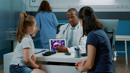 Doctor explaining coronavirus illustration on tablet display to adult and little child in medical cabinet. Man talking about virus spreading animation on device at checkup examination.