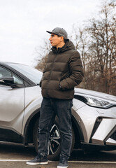 A young cheerful guy in a jacket and a cap stands near a new car