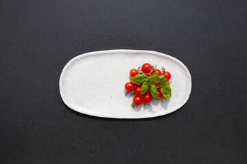 Fresh and tasty cherry tomatoes with basil on white dish on black tabletop. Healthy vegan Food.
