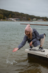 a blonde young girl in a gray coat sits on the parapet by the pond and dips her hand into the water