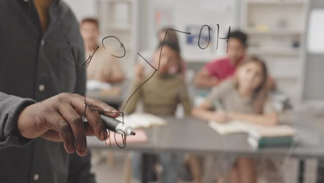 Slowmo closeup of unrecognizable African American teacher drawing structural model of molecular compound with black marker on glassboard explaining material to diverse pupils sitting at their desks