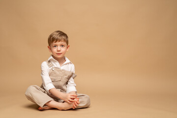 a charming blond four-year-old boy in beige overalls and a white shirt sits on a beige background
