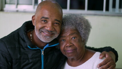A black adult son hugging senior mother posing for camera together