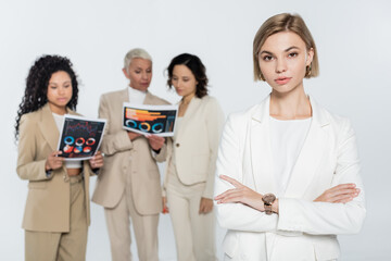 Blonde businesswoman looking at camera near blurred interracial colleagues with papers isolated on grey.