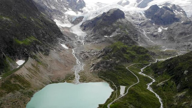 Aerial views with pan down from Stein glacier to heart shaped glacial lake at Sustenpass, Switzerland along with a view of tall peaks of the Swiss Alps