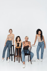 Obraz premium Multiethnic women looking at camera near chairs on grey background.