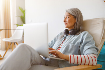 Female person with headphones on her neck looking away and smiling while working on modern notebook