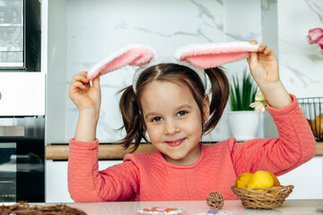 Portrait of a happy dark-haired little girl with bunny ears on her head, sitting at the kitchen table at home.Happy Easter concept.