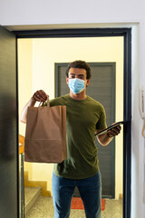 Fototapeta na wymiar Delivery man in front of the apartment with a face mask holding tablet and paper bag