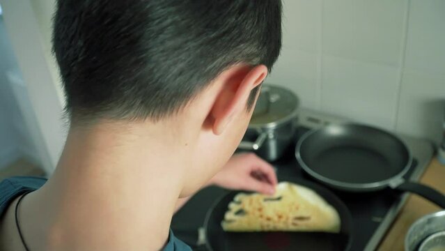 Unrecognizable boy refugee from ukraine cooking pancakes in kitchen of rented hotel room. Concept of self cooking from organic products