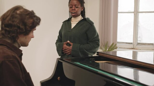 Medium slowmo of young Black woman singing in microphone during vocal practice while her coach playing piano