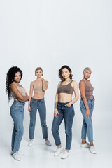Full length of multiethnic friends in jeans and tops posing on grey background.