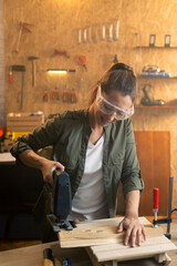 Woman carpenter with safety goggles cutting wood with electric jigsaw in workshop