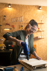 Woman carpenter with safety goggles working with drill in workshop