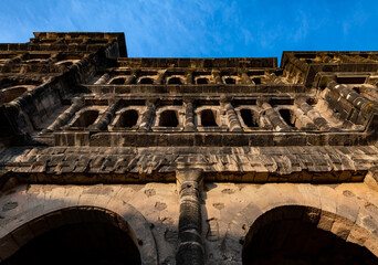 The “Porta Nigra“ (Latin black gate) is a large Roman city gate in Trier, Germany. Historic landmark monument sight in town centre with wheathered sandtone facade, evening light from frog perspective.