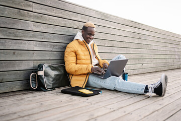 Young trendy African American man sitting outdoors working on laptop smiling. Happy Student...