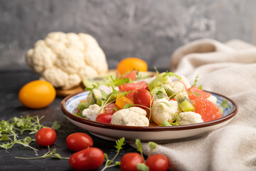 Vegetarian salad of cauliflower cabbage, kiwi, tomatoes, microgreen sprouts on black concrete background. Side view, selective focus.