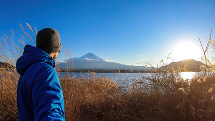 Man walking through golden grass at the side of Kawaguchiko Lake, Japan with the view on Mt Fuji, while taking a selfie. The mountain surrounded by clouds. Serenity and calmness. Exploring new places