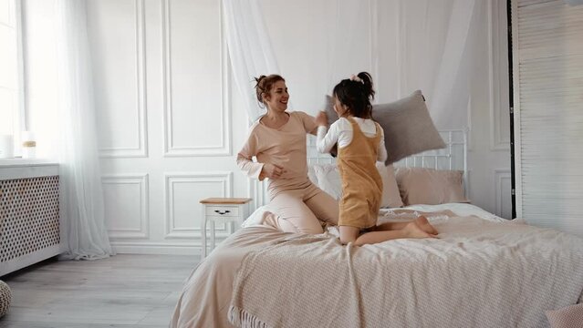 Happy family young mom baby sitter and little kid daughter enjoy funny pillow fight on bed, carefree mother nanny laughing playing game together having fun with child girl in bedroom in the morning