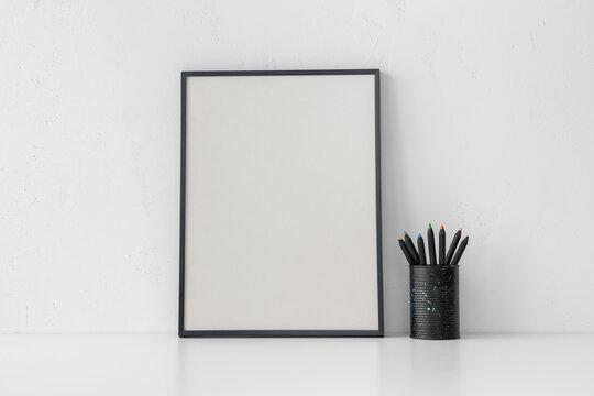 Black picture frame and pencils in metal cup on white wall. Stylish home decor design. Mockup..