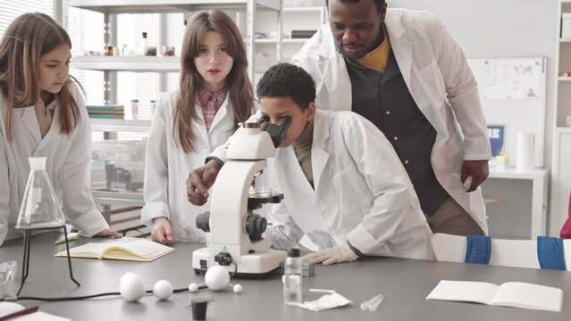 Medium slowmo of group of multiethnic middle school pupils and their African American male teacher wearing white lab coats using microscope while doing experiments at lesson in Science class