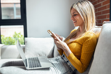 Beautiful young woman working with her laptop while using mobile phone sitting on a couch at home.