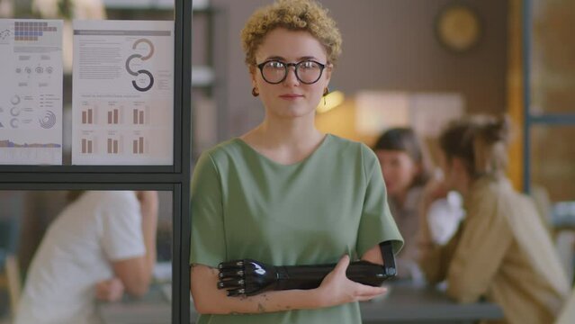 Portrait of young Caucasian businesswoman with prosthetic arm standing by glass wall in office and posing on camera