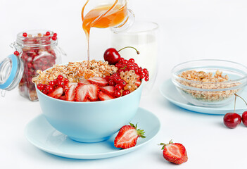 Pouring honey into blue granola bowl with fresh berries on white background. Healthy food