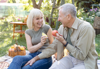 Laughing senior spouses sitting on field grass and eating fruits, talking and smiling, resting outdoors in their garden