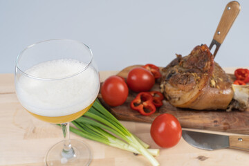 A glass of beer, a fork stuck in a pork knuckle and a knife next to it on a cutting board, meat cooked in the oven with vegetables