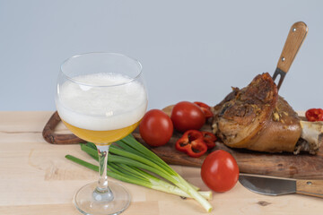 A glass of beer, a fork stuck in a pork knuckle and a knife next to it on a cutting board, meat cooked in the oven with vegetables