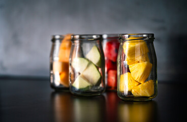 Variety small pieces of tropical fruits in cleared bottles on black table, each kind of fruit is separated into a glass bottle as snacks, blurred background grey wall. Dark food style photography